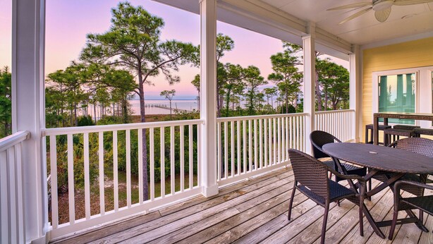 Un espace extérieur abrité, équipé d’une table et de chaises, d’un ventilateur de plafond, et offrant une vue sur des arbres et une plage.