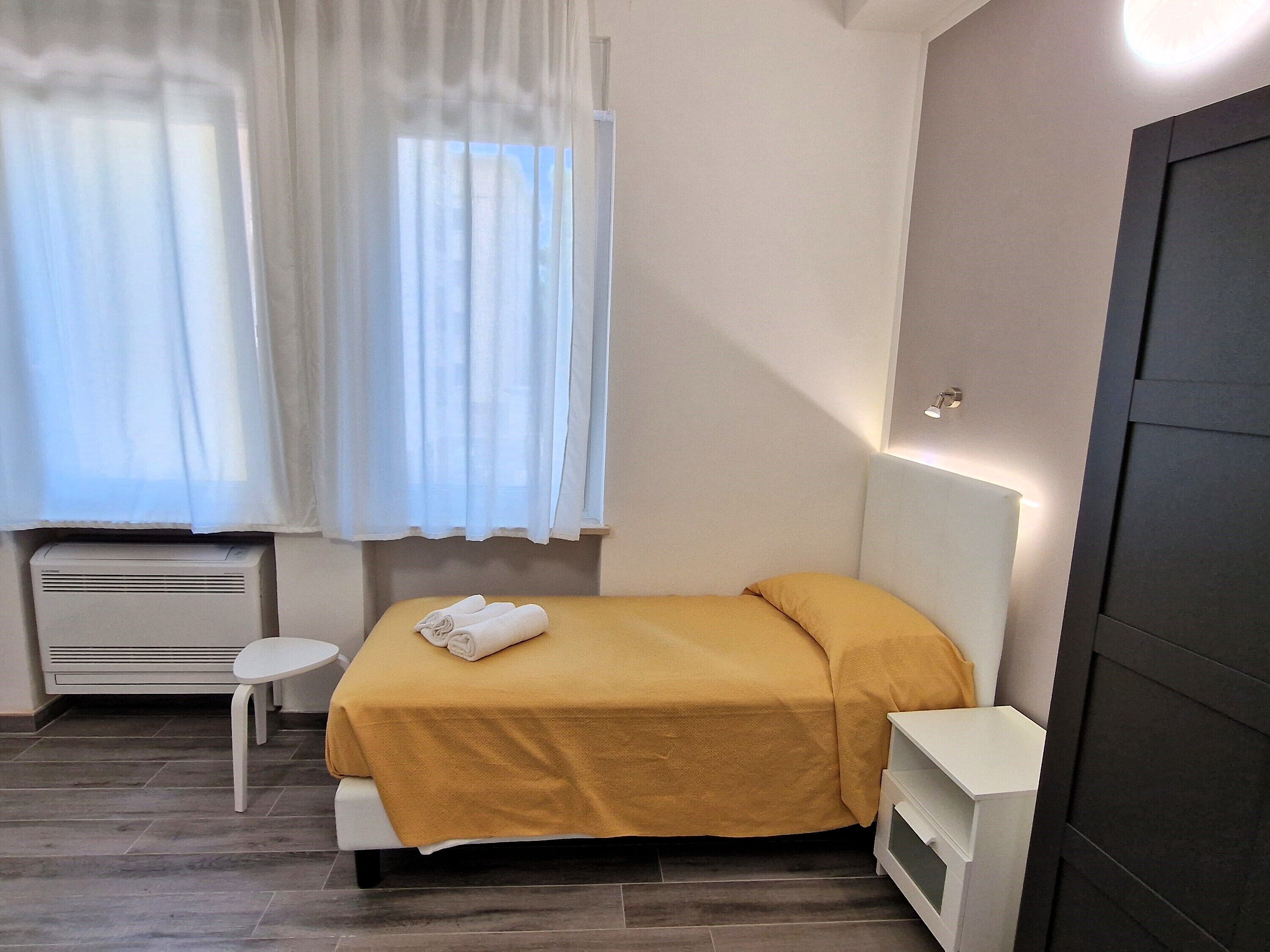 Ein kleines, aufgeräumtes Schlafzimmer mit einem Bett, einem Nachttisch, einem kleinen Hocker und einem Heizgerät.