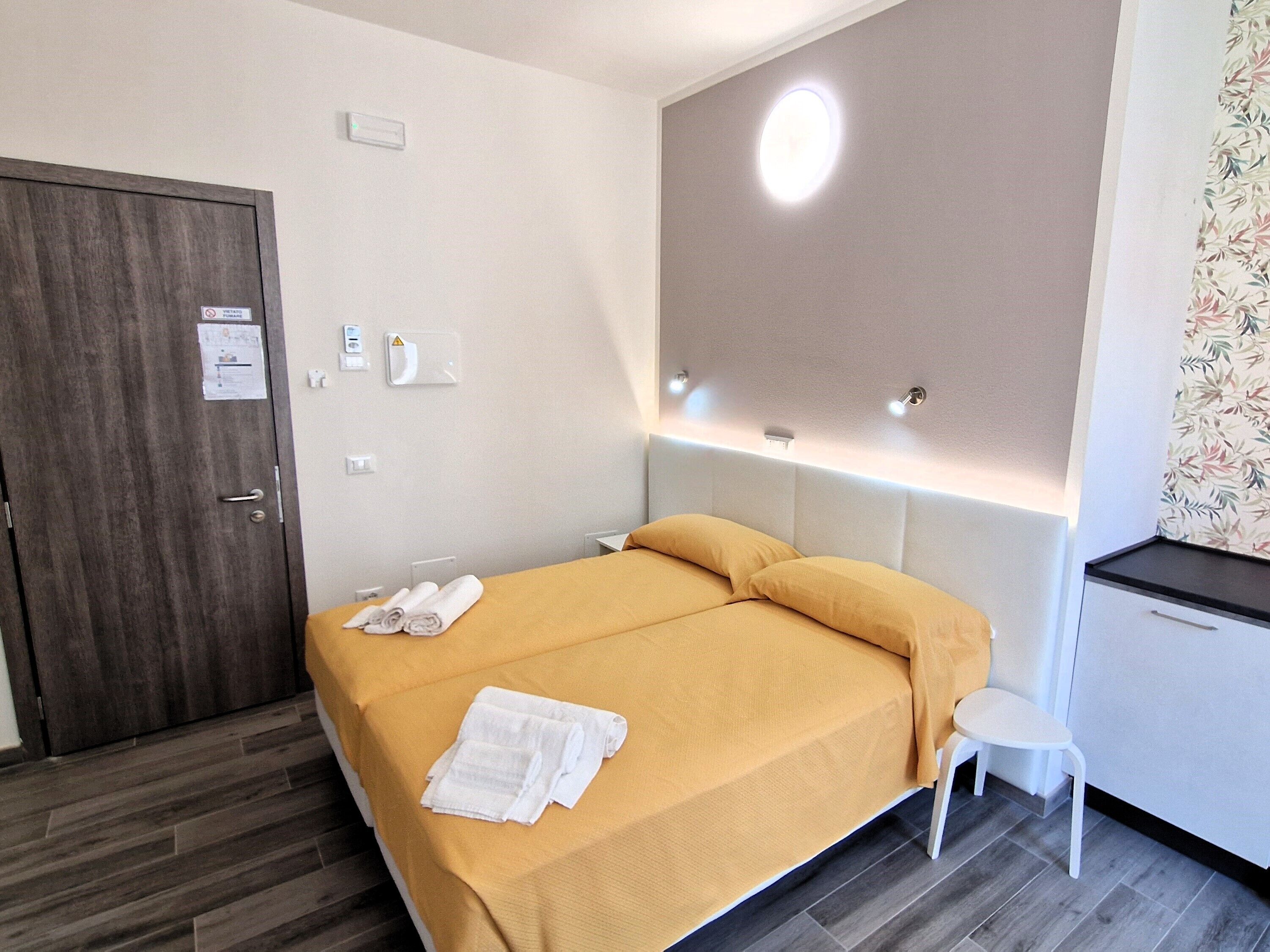 Una habitación de hotel con una cama, un taburete blanco pequeño, una puerta y una lámpara fijada a la pared.