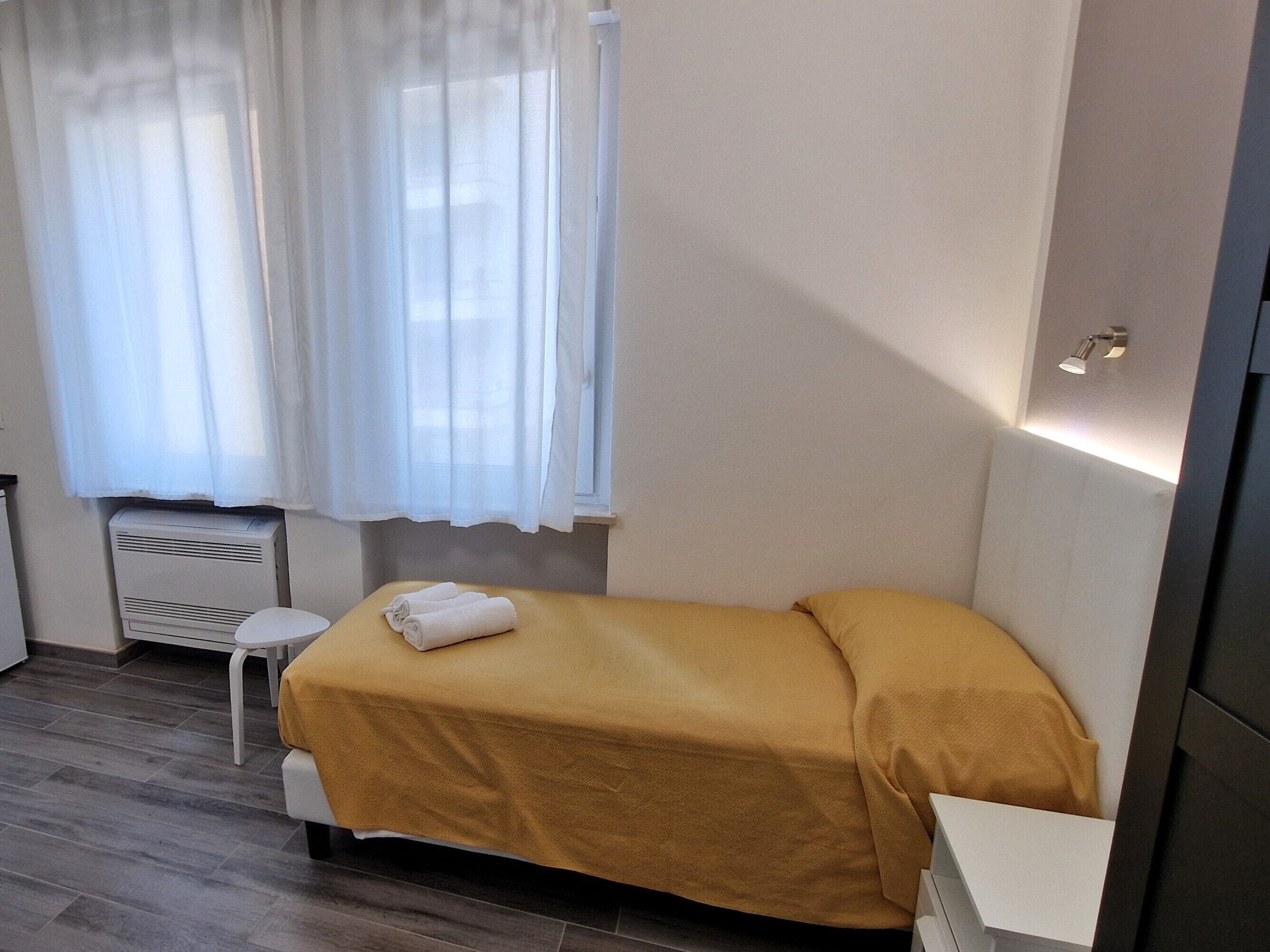 A small, single-bed room with a yellow bedspread, a white chair, a small table, and a window with white curtains.