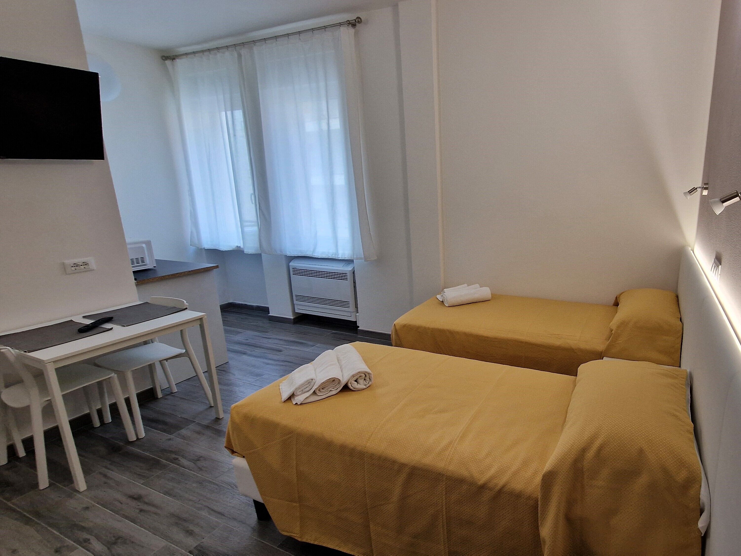 A hotel room with two single beds, a desk with a chair, a TV mounted on the wall, and a window with white curtains.