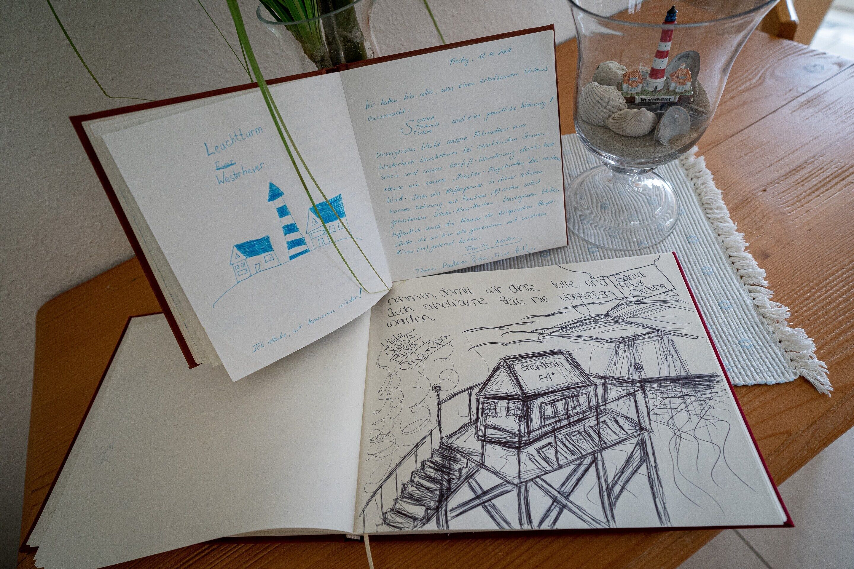Un cuaderno abierto con bocetos y texto, un florero de vidrio con rocas y una maqueta de faro, y una mesa de madera.