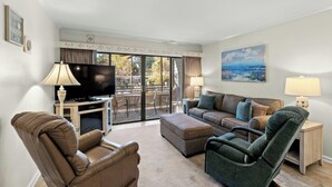 A spacious living room with a flat-screen TV, comfortable seating, a balcony with a view, and a painting on the wall.