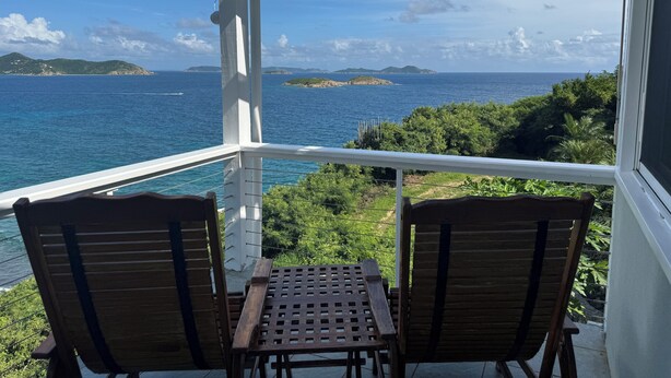 Ein Balkon mit zwei Holzstühlen und einem kleinen Tisch, der Blick auf die Küste mit klarem, blauem Wasser und entfernten Inseln bietet.