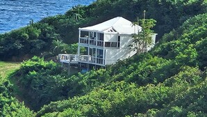 Ein weißes, zweistöckiges Haus mit Balkon, umgeben von üppiger Vegetation und mit Blick auf ein Gewässer.