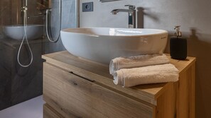 Un baño con un lavamanos grande tipo recipiente, un cabezal de ducha manual, una botella de champú y un mueble de tocador de madera.