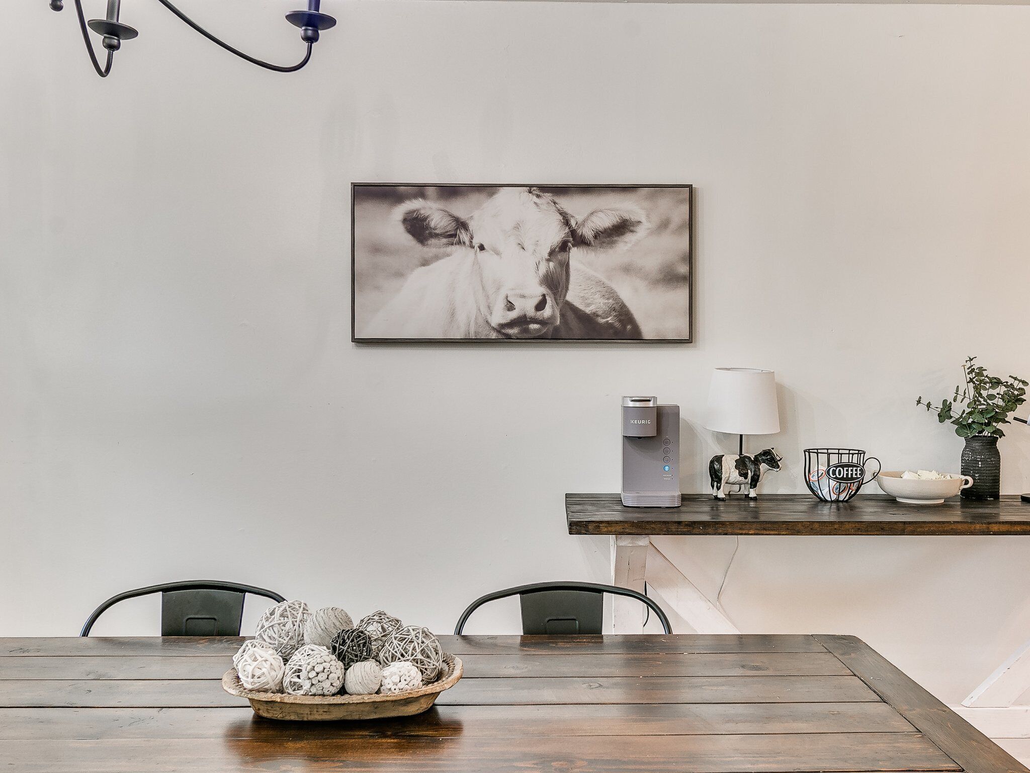 Een eetgedeelte met een houten tafel, stoelen, een schaal decoratieve items, een waterdispenser, een koffiekop, een lamp en een lijst met een afbeelding van een koe aan de muur.