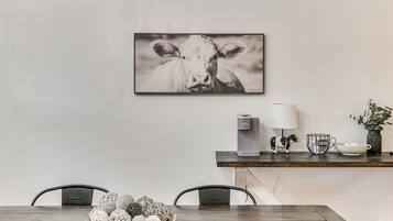 Een eetgedeelte met een houten tafel, stoelen, een schaal decoratieve items, een waterdispenser, een koffiekop, een lamp en een lijst met een afbeelding van een koe aan de muur.