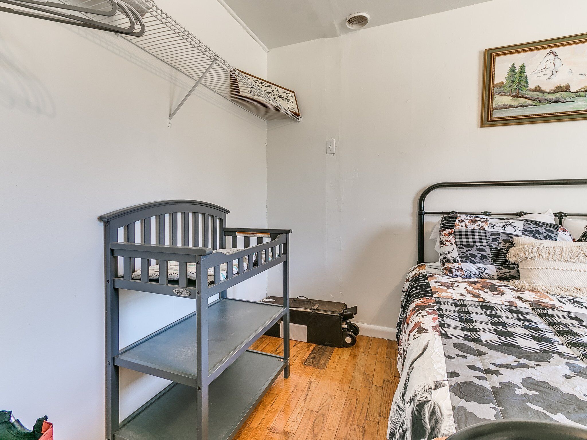 A bedroom with a bunk bed, a bed with patterned bedding, a painting on the wall, and a suitcase on the floor.