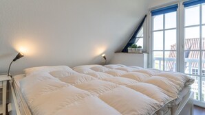 Un letto rifatto con biancheria bianca in una stanza dalle pareti bianche e soffitto spiovente. Sono presenti due lampade da comodino e una finestra con tende blu.
