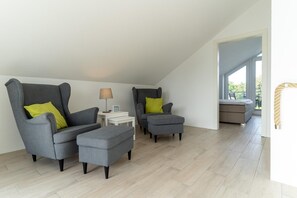 A modern loft bedroom with a high ceiling, grey upholstered armchairs, a small white nightstand, and a grey ottoman.