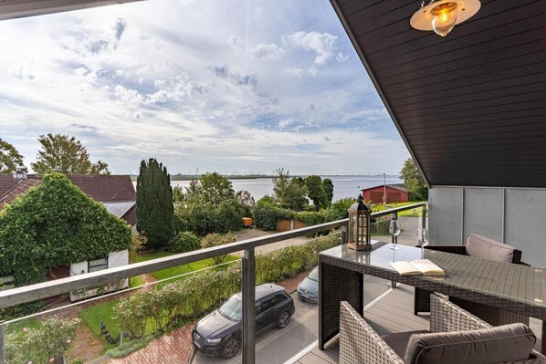 A balcony with a view of a lake, a car parked outside, and a table with two wine glasses and a book.