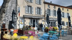 An outdoor café with tables and chairs, a building with shutters, and people walking by. - Our establishment is located not far from Clermont-Ferrand in the charming littl (Saint-Saturnin)