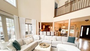 A spacious living room with a large L-shaped sectional sofa, a coffee table, and a view of the kitchen and dining area.