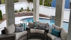 A covered outdoor seating area with wicker furniture and cushions, a pool in the background, and a white fence.