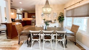 A spacious dining area with a large wooden table and white chairs, a chandelier, and a view of the kitchen in the background.