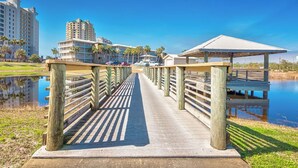 Un muelle de madera que lleva a una masa de agua, con edificios y palmeras al fondo.