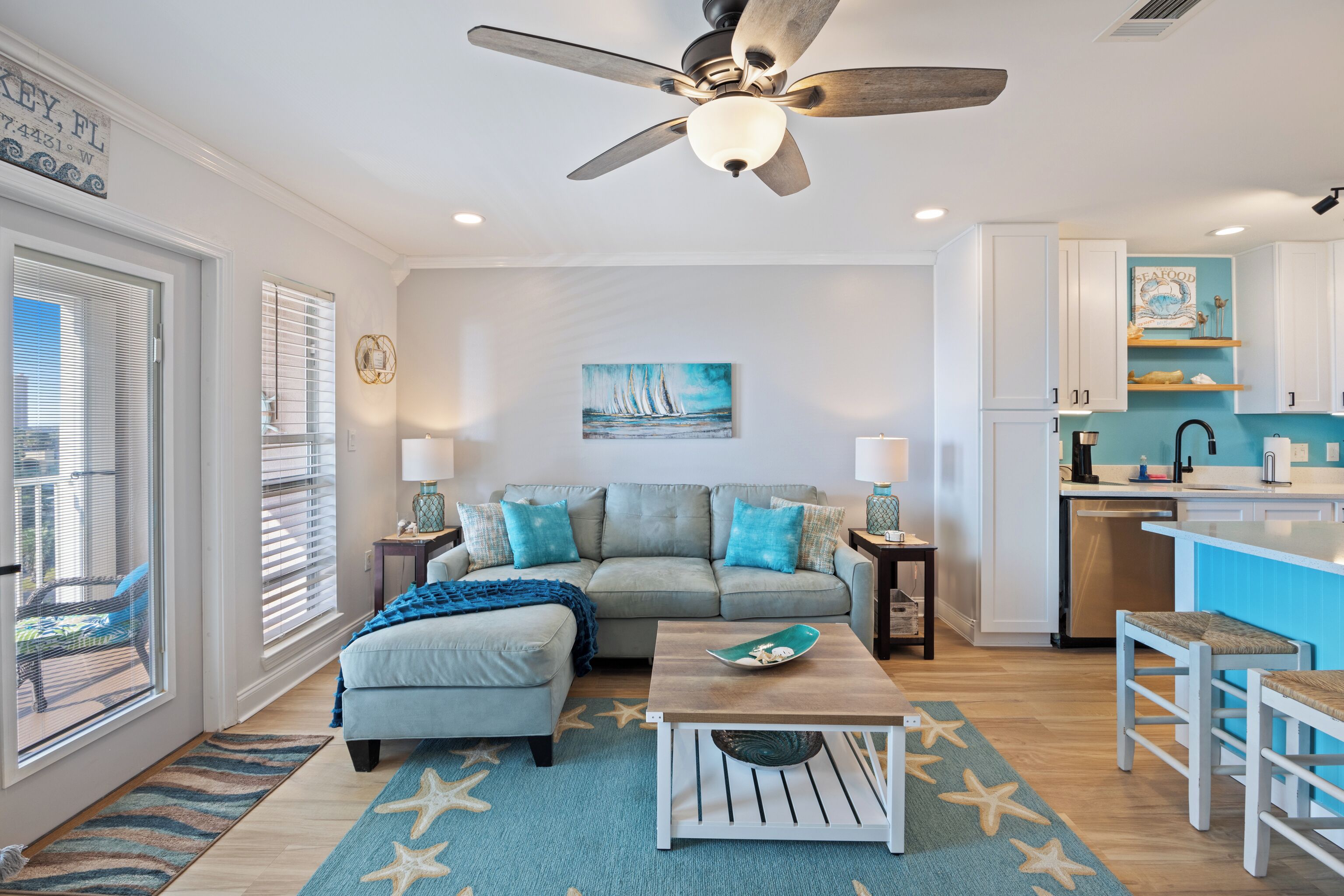 A living room with a sofa, coffee table, and starfish rug. There is a kitchen area with white cabinets and a blue backsplash.