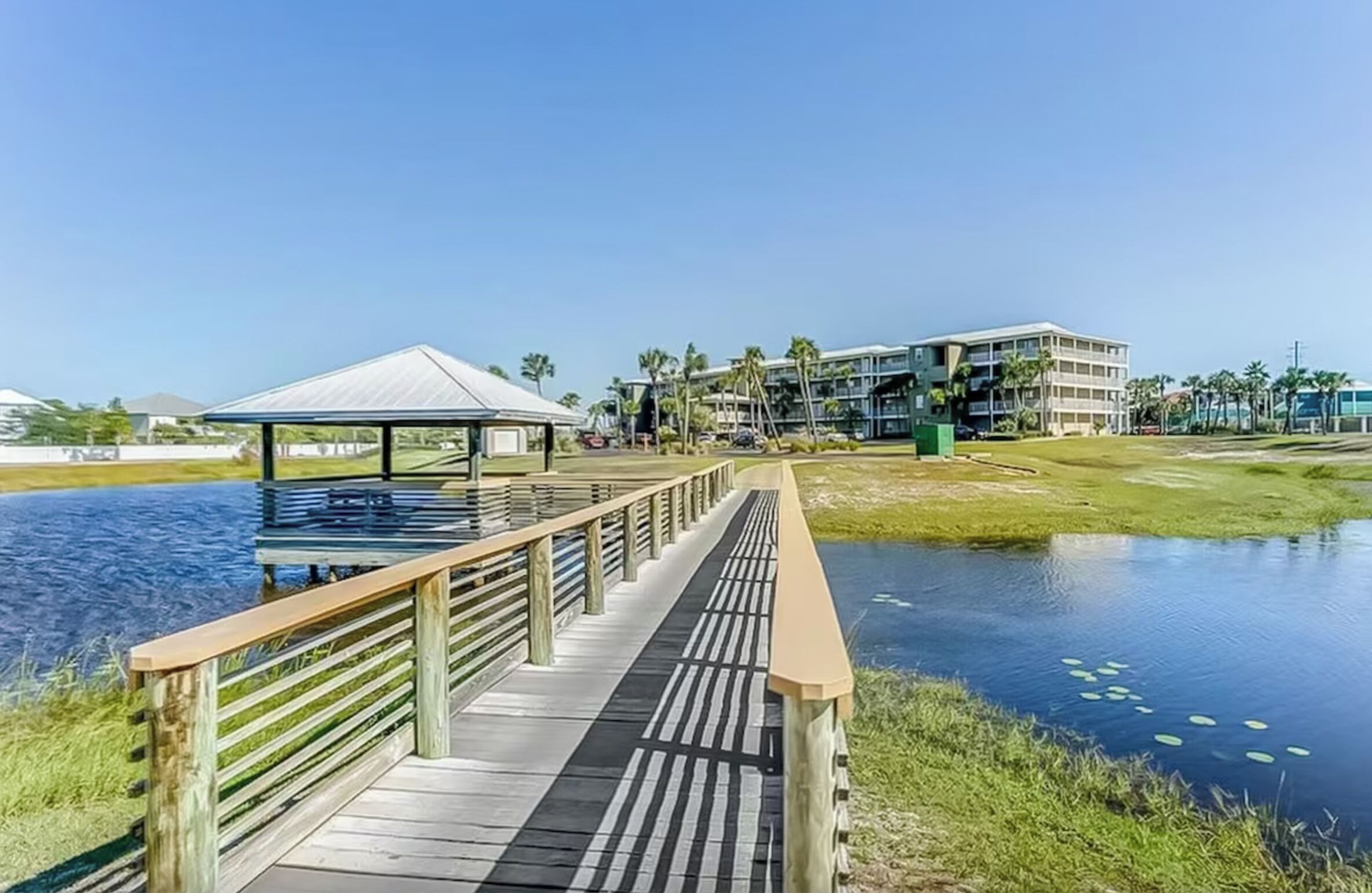 A wooden boardwalk leading to a gazebo by a body of water, with a building in the background.