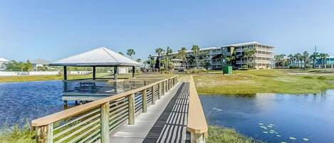 A wooden boardwalk leading to a gazebo by a body of water, with a building in the background.