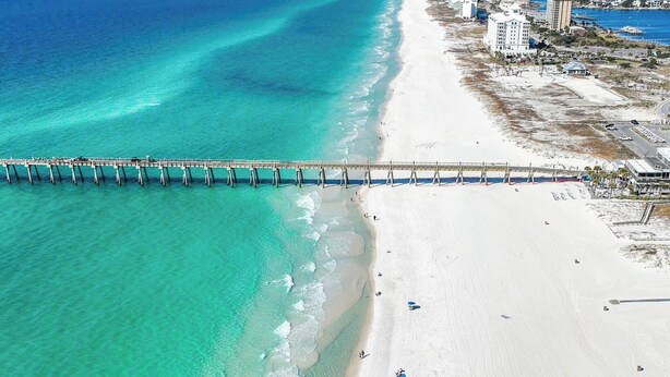 Una playa con un muelle que se adentra en aguas turquesas, una orilla arenosa y edificios al fondo.