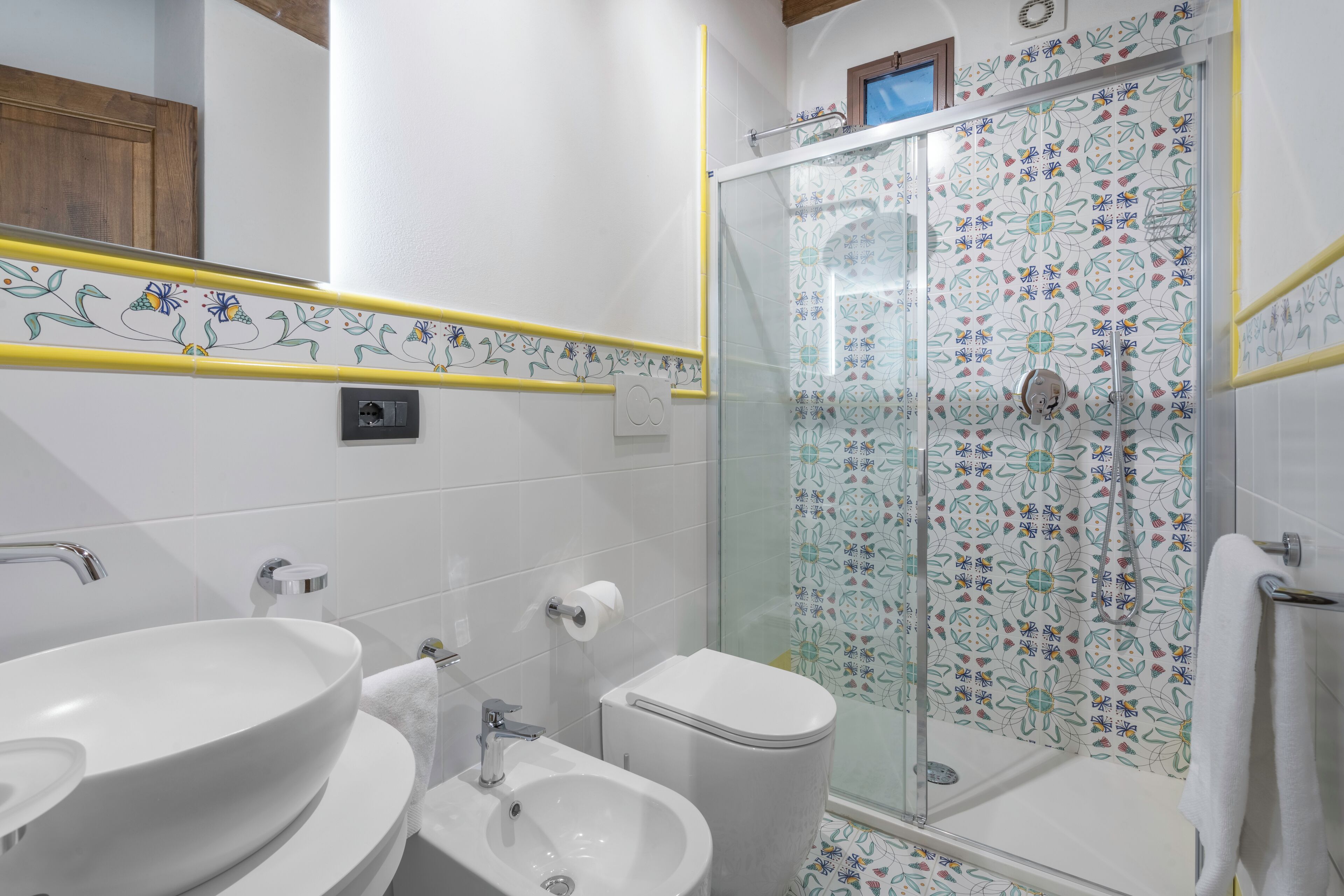 A bathroom with a large white sink, a bidet, and a toilet. There is a glass shower enclosure with a floral tile pattern.