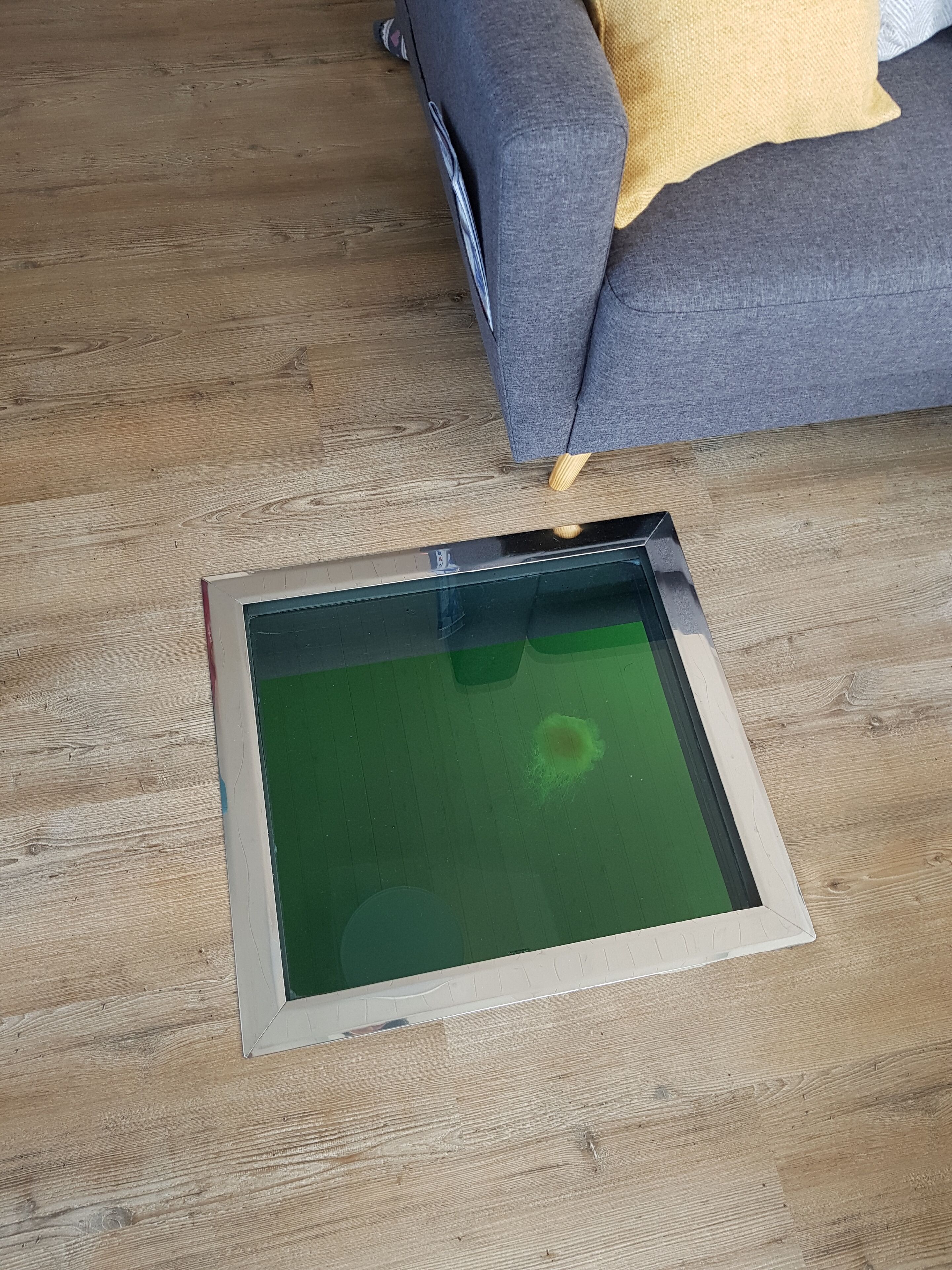 Um tablet sobre um chão de madeira, com uma tela verde, um sofá cinzento com uma almofada amarela, e uma mesa de madeira ao fundo.