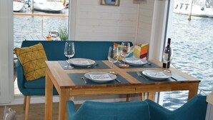 A dining table set with plates, glasses, and cutlery, a wooden bench with a cushion, and a view of the water through a window. - Houseboat Hecht with spacious roof terrace in Schleswig (Schleswig)