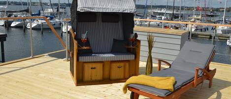 Una terraza de madera con una cabaña pequeña, una tumbona y vistas al puerto deportivo con barcos.