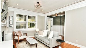 Una sala de estar con un sofá gris, una silla de madera y una mesa de centro. Hay una ventana con persianas, una lámpara y un candelabro.