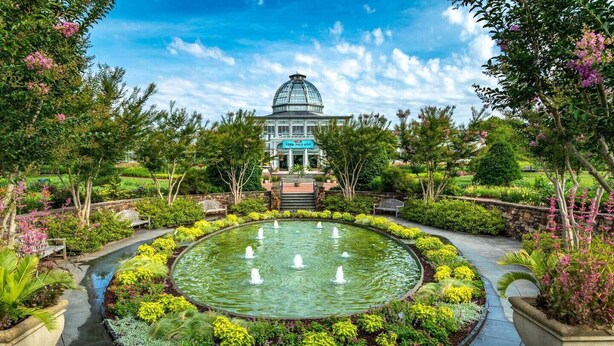 Una gran fuente circular rodeada por un jardín con diversas plantas y un edificio con cúpula al fondo.