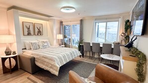 A modern hotel room with a large bed, a sitting area with a table and chairs, a TV mounted on the wall, and a view of the city through the windows. - Lux 1BR Ski IN/OUT: King+Queen, No Sleeper! @Lift (Snowshoe)