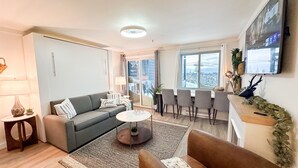 A modern living room with a grey sofa, a brown armchair, a coffee table, and a dining area with four chairs. A TV is mounted on the wall, and there's a window with a view of a snowy landscape. - Lux 1BR Ski IN/OUT: King+Queen, No Sleeper! @Lift (Snowshoe)