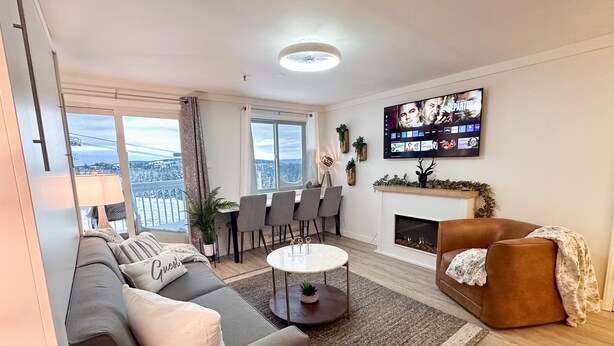 A modern living room with a sofa, a coffee table, and a fireplace. There is a dining area with chairs and a TV mounted on the wall. - Lux 1BR Ski IN/OUT: King+Queen, No Sleeper! @Lift (Snowshoe)