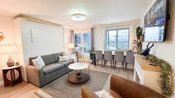 A modern living room with a grey sofa, a brown armchair, a coffee table, and a dining area with four chairs. A TV is mounted on the wall, and there's a window with a view of a snowy landscape.