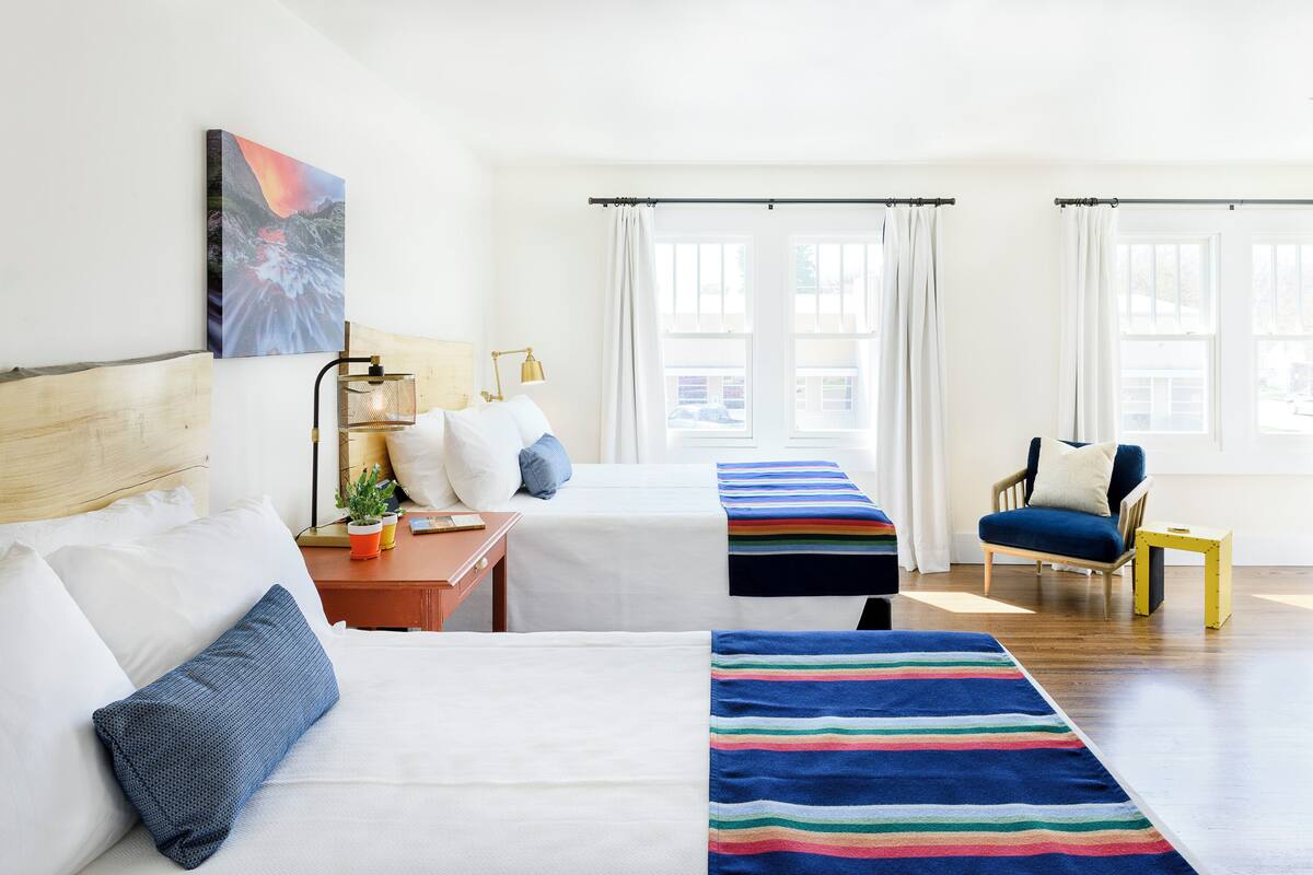 Une chambre d’hôtel avec deux lits, une chaise, une petite table avec une lampe et un tableau au mur.
