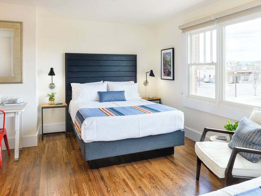 A modern hotel room with a large bed, a desk, a chair, and a window with a view of buildings.