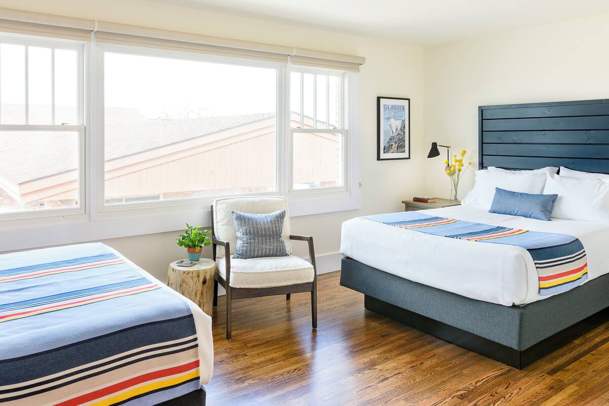 Une chambre à coucher comprenant un grand lit, un lit plus petit, une chaise, une table de chevet, une lampe et une fenêtre donnant sur des immeubles.