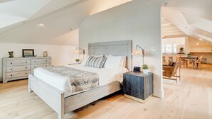 A spacious loft bedroom with a large bed, wooden nightstands, and a dining area visible through an open door. - Sherman Lodge (Kalispell)
