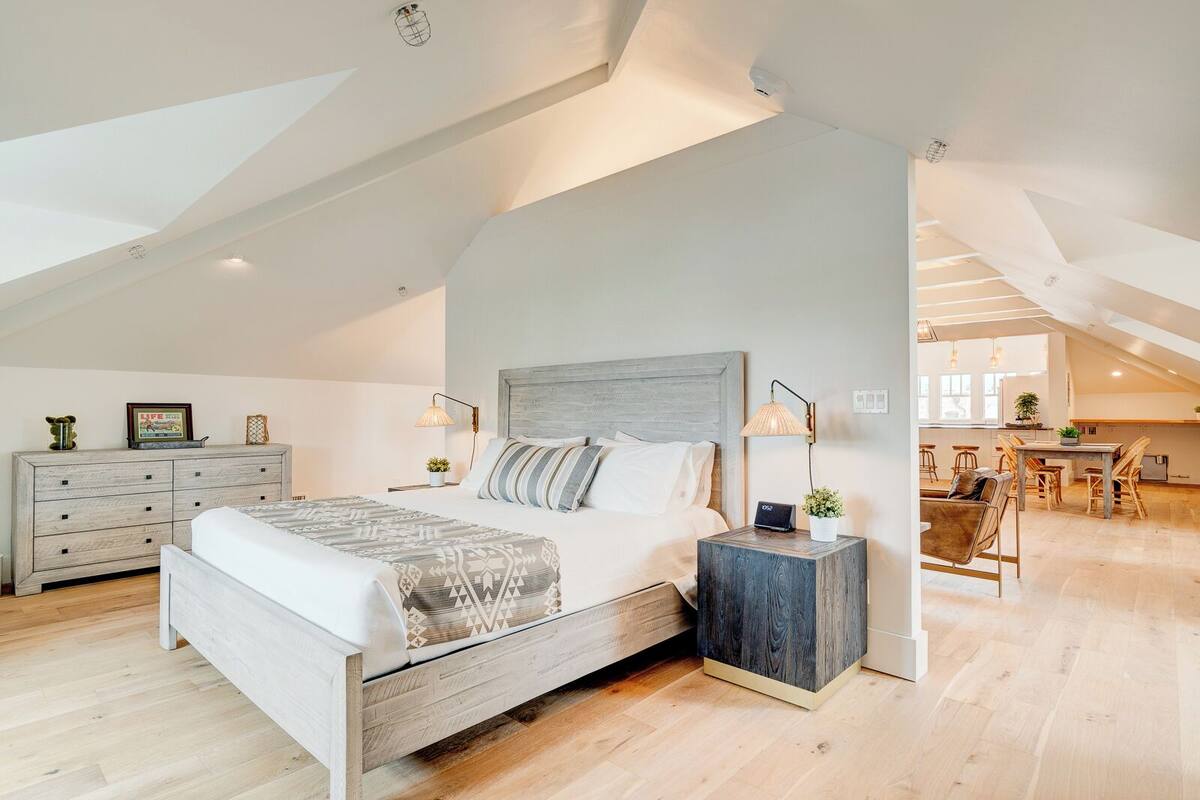 Une chambre spacieuse aménagée en loft, avec un grand lit, des tables de chevet en bois et un coin repas visible à travers une porte ouverte.