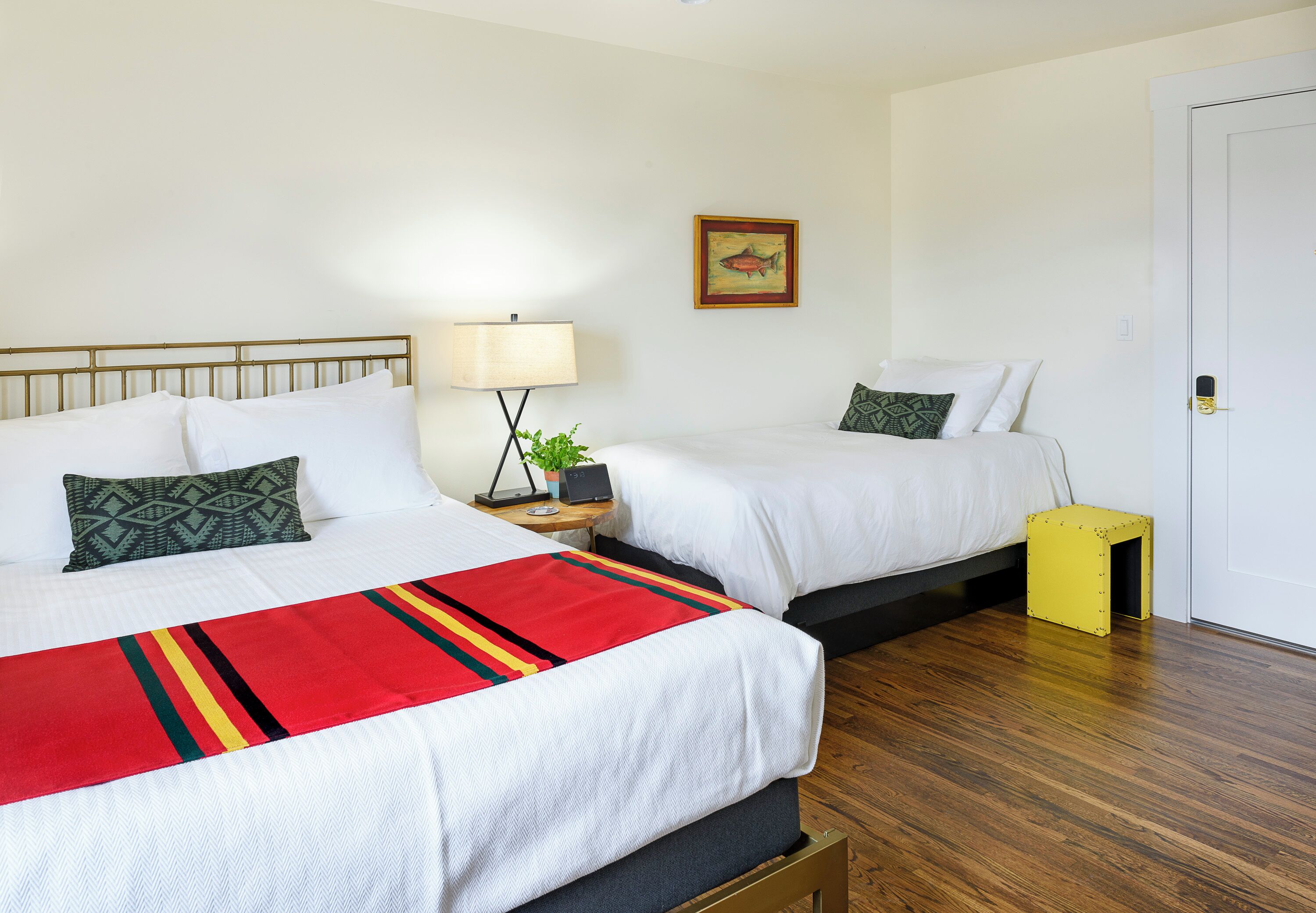 A hotel room with two beds, a bedside table with a lamp, a small plant, and a door.