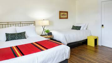 A hotel room with two beds, a bedside table with a lamp, a small plant, and a door.