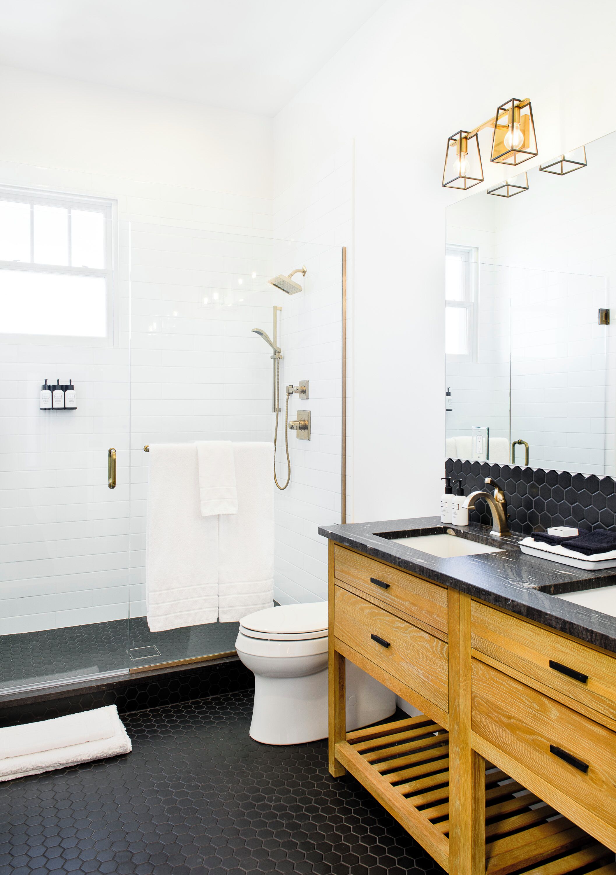 A modern bathroom with a black and white tiled floor, a wooden vanity with a black countertop, a white toilet, a shower with a handheld showerhead, and a large mirror above the sink.