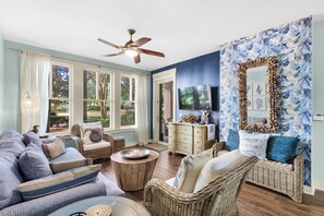 A living room with a large window, a ceiling fan, wicker furniture, a wooden coffee table, and a wall-mounted TV.