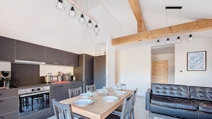 A modern kitchen with dark cabinets, a wooden dining table set for six, and a black leather sofa in the living area. - Snow Roc - 6/8 persons - Ski to door access - Mountain view (Saint-Jean-d´Aulps)