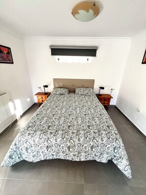 Un dormitorio con una cama grande, cabecera, mesitas de noche y ventana con persianas.