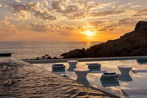 Ein Pool ohne Ende mit weißen Liegestühlen, eine felsige Küstenlinie und ein Sonnenuntergang über dem Meer.