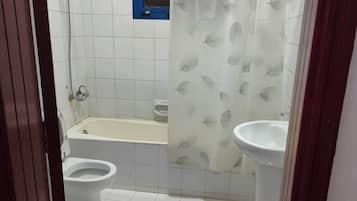 A bathroom with a bathtub, two toilets, a shower curtain, and a window with a blue frame.