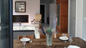 Un comedor con una mesa de madera dispuesta para cuatro personas, rodeada de sillas de madera. Un candelabro cuelga del techo, y un espejo está colgado en la pared.