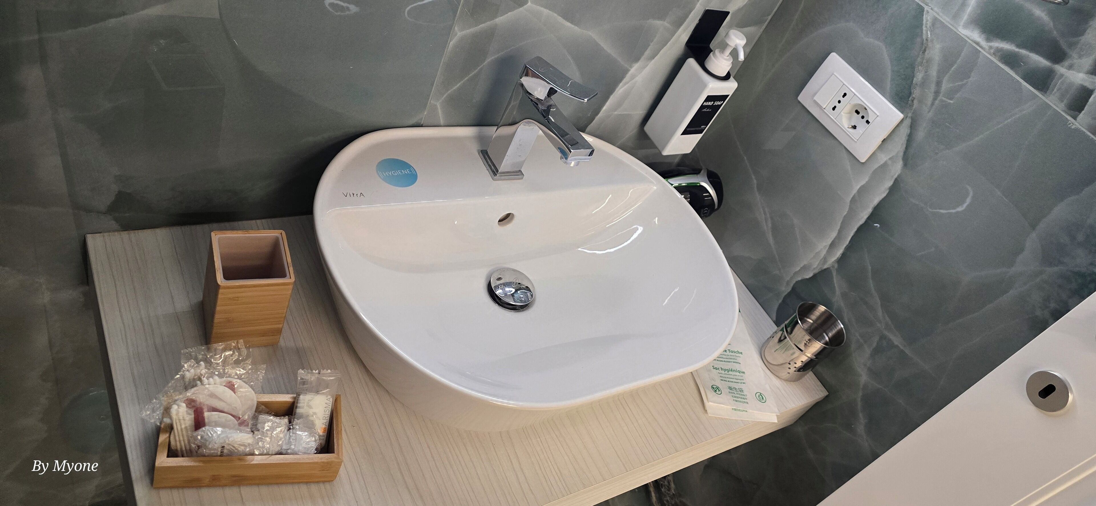 Kylpyhuone, jossa on valkoinen pesuallas, peili, saippua-annostelija ja pieni puinen alusta, jossa on hygieniatuotteita.